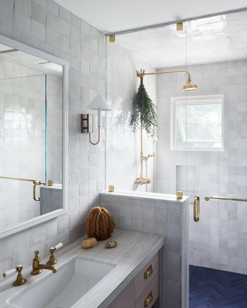 Bathroom with floor-to-ceiling white zellige tile, unlacquered brass faucet and shower fixtures, navy herringbone floor tile and hanging eucalyptus
