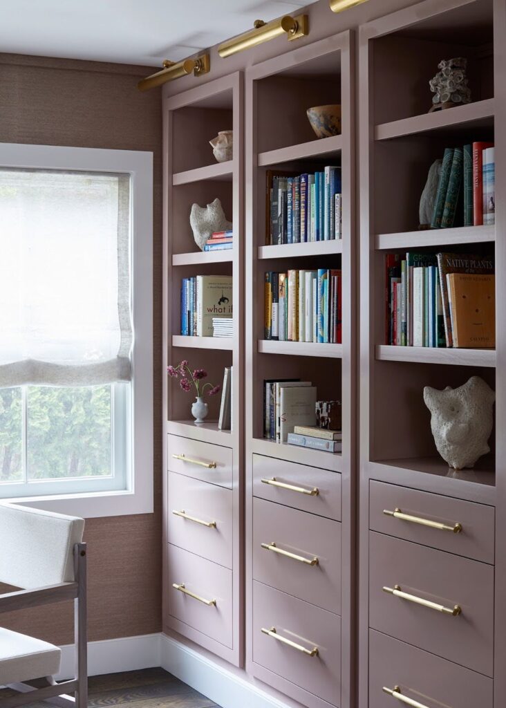  Home office with custom floor-to-ceiling dusty rose lacquered bookshelves with brass hardware against warm brown grasscloth wallpaper and linen roman shade
