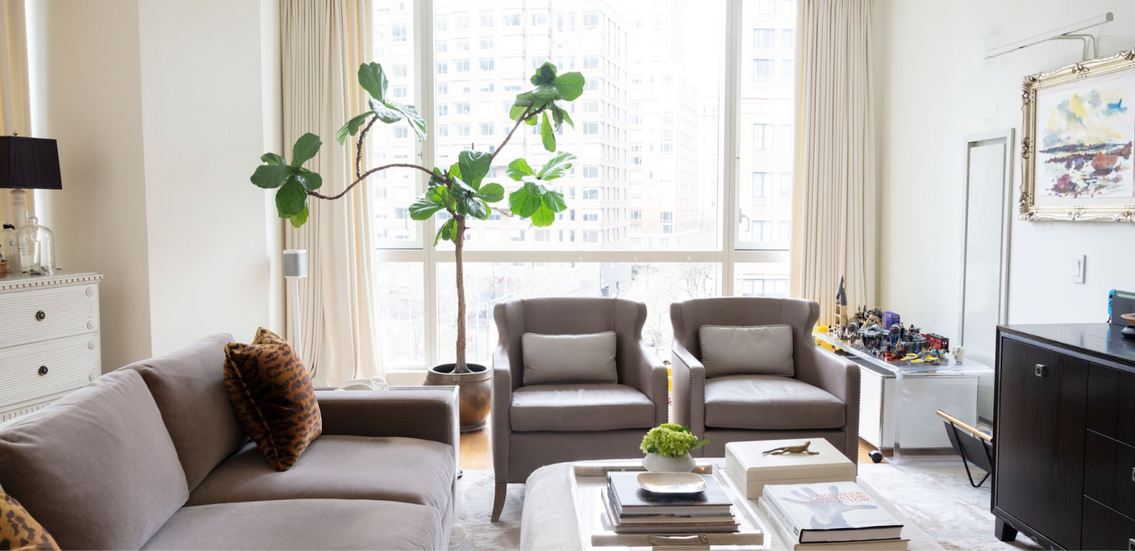 Bright New York City apartment living room with warm greige sofa and armchairs, cream drapery panels, fiddle leaf fig tree and colorful artwork in gilded frame