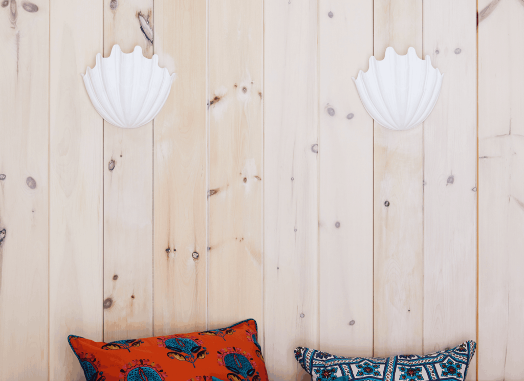 Two white scalloped shell-shaped wall sconces flank patterned throw pillows in orange and navy blue geometric prints on a light wood shiplap wall, showcasing sculptural lighting and pattern layering in a coastal pool house.