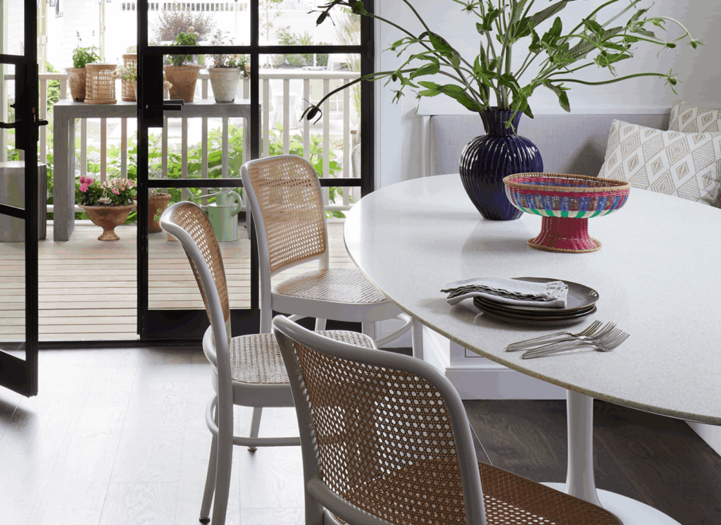 A round white dining table with woven rattan chairs sits in a bright room with floor-to-ceiling windows and black frames opening onto a wooden deck, with natural fiber placemats and accessories on the table surface.