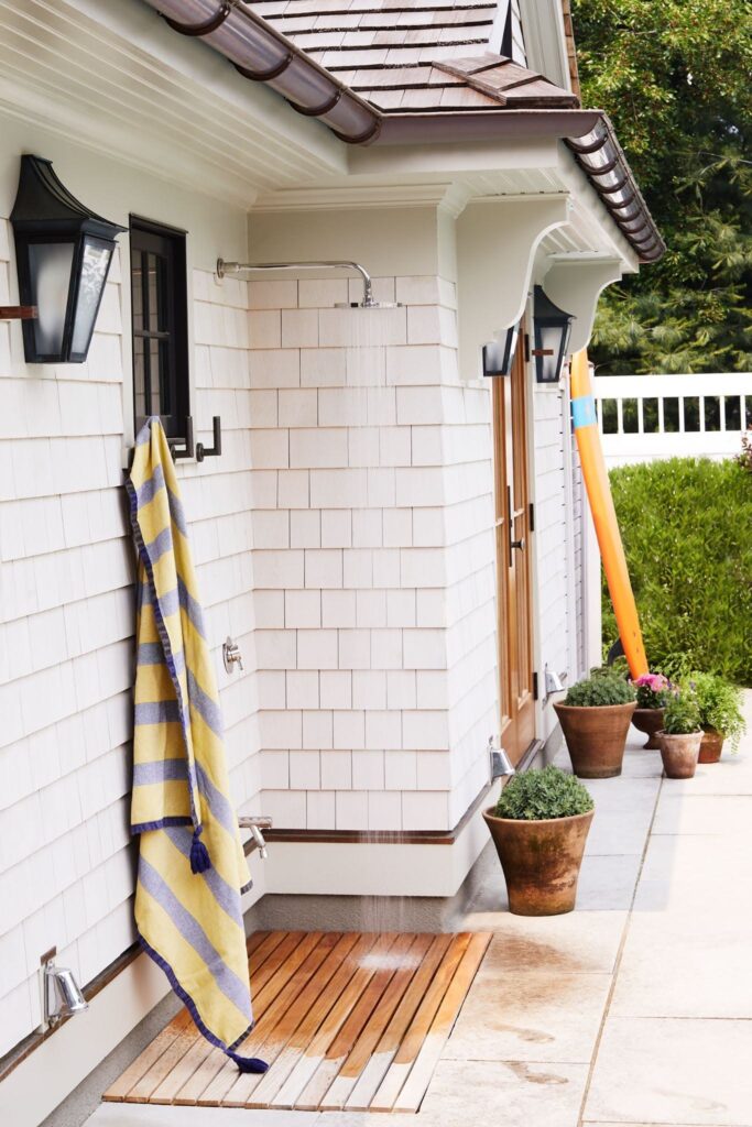 An open-air outdoor shower features wooden decking underfoot, quality brass fixtures, and yellow-and-navy striped towel hanging on the exterior, with white shingle siding and potted plants visible beyond.