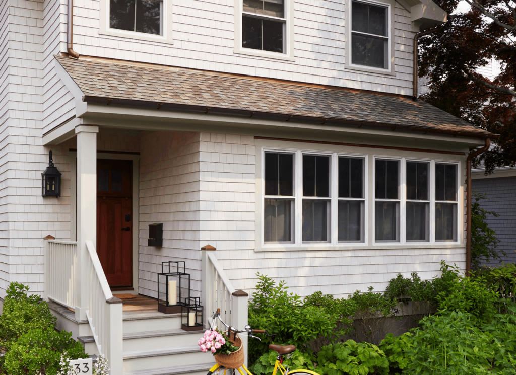A cottage exterior features white cedar shingles, a burgundy red front door with black hardware and lantern fixture, a shallow porch with white railings.