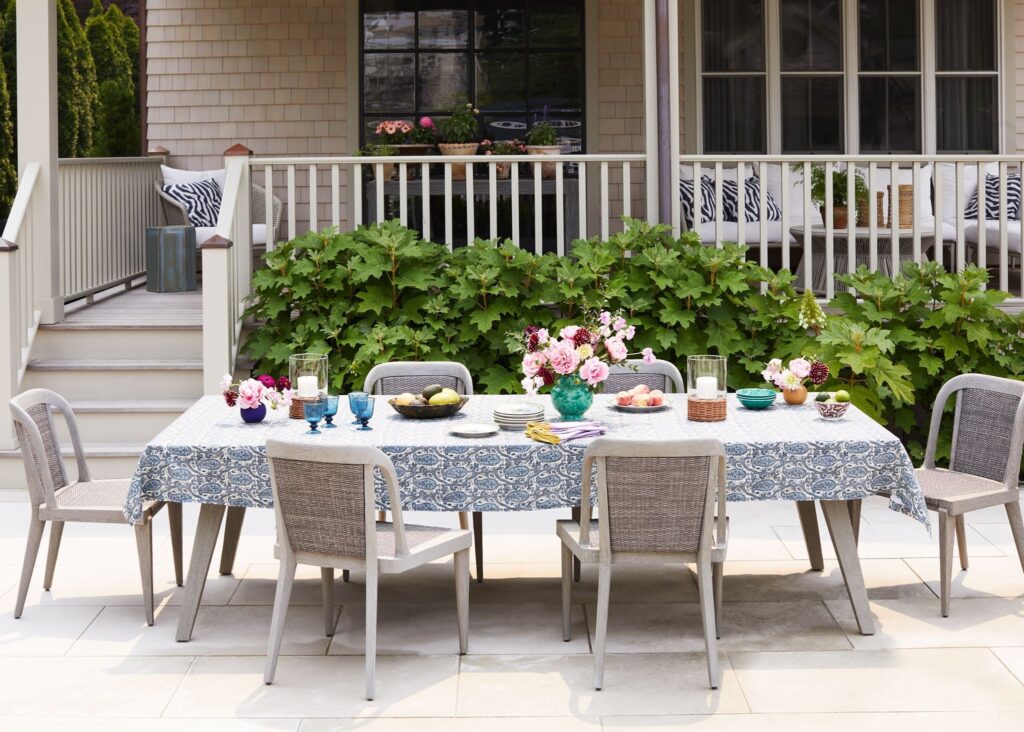 A white outdoor dining table draped with a navy and white patterned tablecloth is set with plates and glasses, surrounded by woven rattan chairs, fresh flowers in teal and gold vessels, flowering shrubs, and a deck railing with zebra-printed pillows visible in the background.