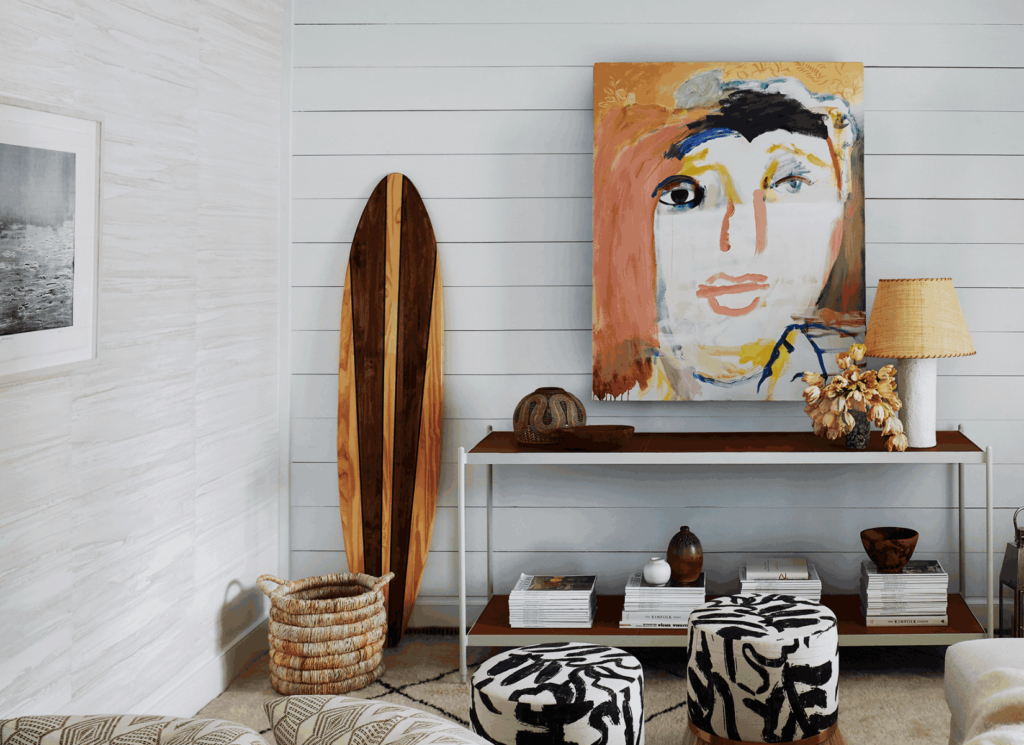 Vibrant contemporary portrait artwork in warm and cool tones hangs above a two-tier console table styled with white art books, sculptural ceramics, and a rattan basket, demonstrating how to layer art and accessories in a coastal living space.
