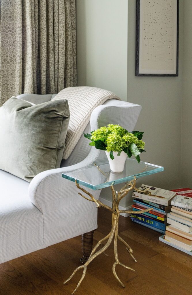 Reading nook detail with patterned cornice and drapery, a branch-form glass side table, a green velvet pillow and a stack of books including Infinite Jest