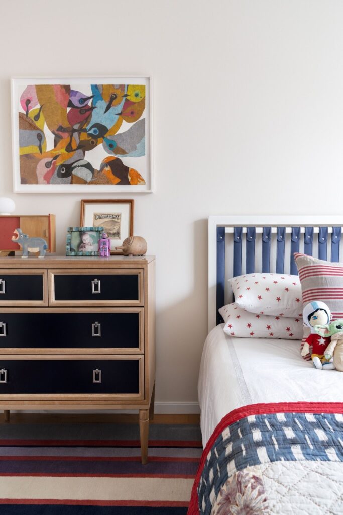 Child's bedroom with a navy leather tab-top headboard, a colorful bird artwork, star print pillows and a mix of vintage and handmade objects on a wooden dresser