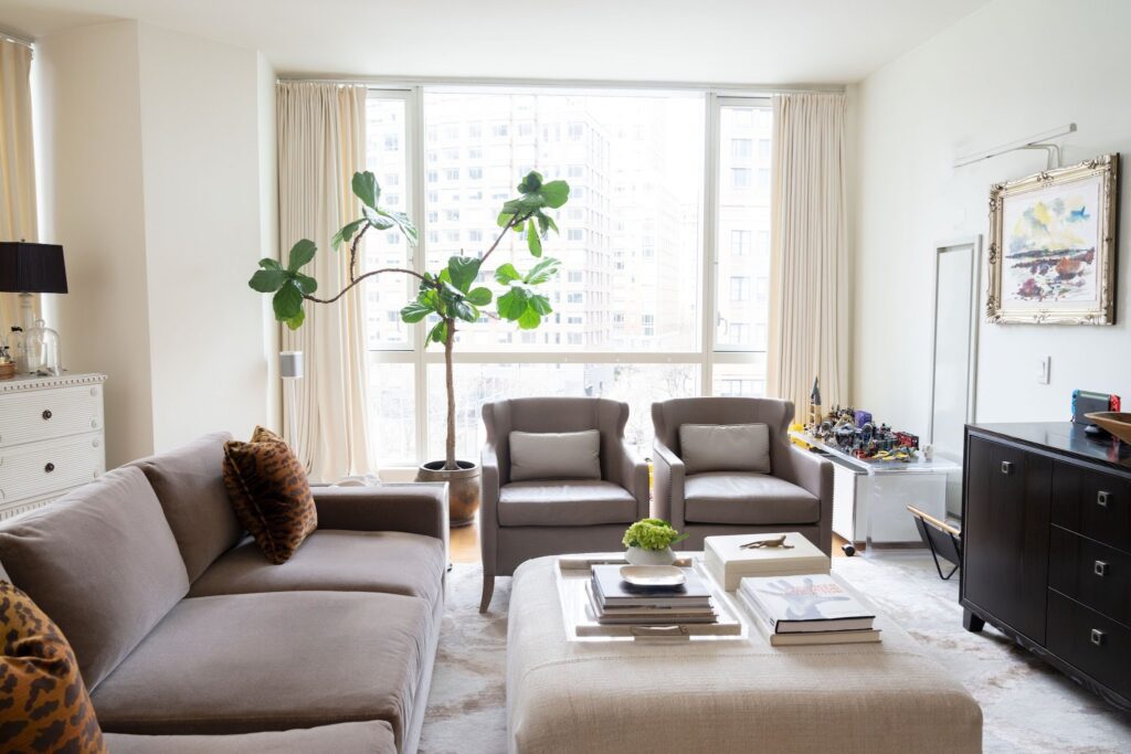 Living room vignette pairing an oversized fiddle leaf fig with a small gilt-framed landscape painting, illustrating contrasting scale in a high-ceilinged Manhattan apartment