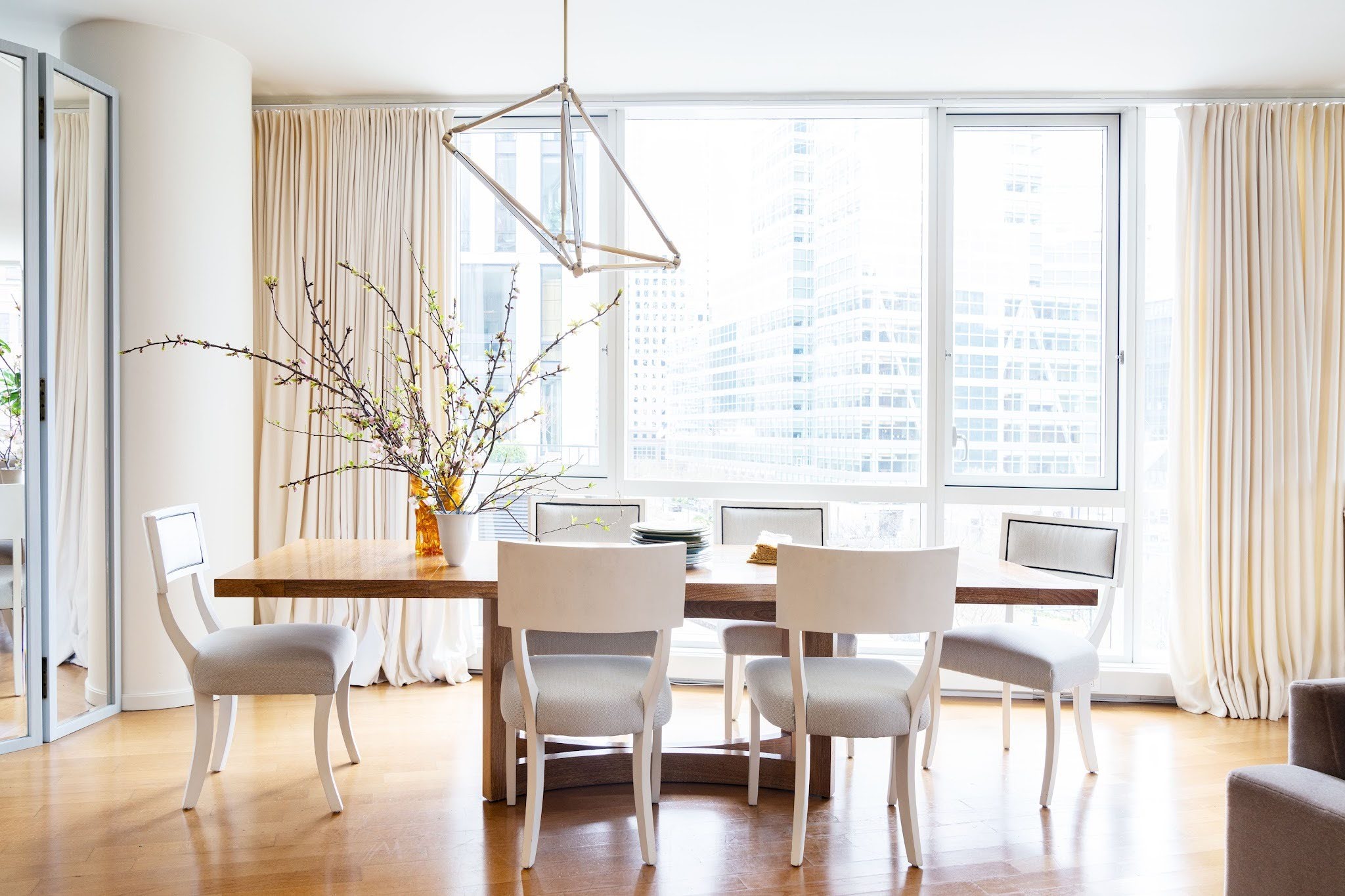 Alt: Open-plan Tribeca dining area with a live-edge wood table, ivory dining chairs and a geometric wood pendant light framed by floor-to-ceiling windows overlooking the Manhattan skyline